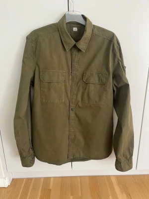 Olivgrön overshirt från C.P. Company - Olivgrön overshirt från C.P. Company med två bröstfickor och klassisk krage. Långärmad modell i bomull med den ikoniska linsen på ärmen. Stängs med knappar framtill och har en clean, modern look.
