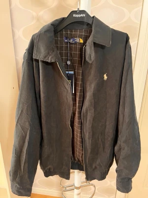Svart jacka från Polo Ralph Lauren - Stilren svart jacka från Polo Ralph Lauren med klassisk krage och dragkedja. Jackan har rutigt innerfoder och den ikoniska loggan broderad på bröstet. Perfekt för dig som vill ha en tidlös look med snygga detaljer. passar som en XL