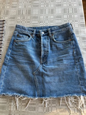 Blå jeanskjol med fransad kant - Säljer en jeanskjol i kort modell med fransad nederkant. Kjolen har knappgylf och har storlek 36. 