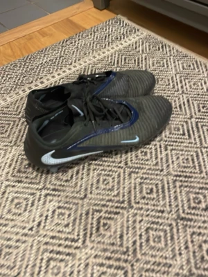 Svarta Nike fotbollsskor med blå detaljer - Säljer ett par svarta Nike fotbollsskor med blå och vita detaljer. Skorna har snörning och en låg profil, samt mönstrad ovansida för bättre bollkontroll. Perfekta för dig som vill ha stilrena och funktionella skor på planen. Priset kan diskuteras