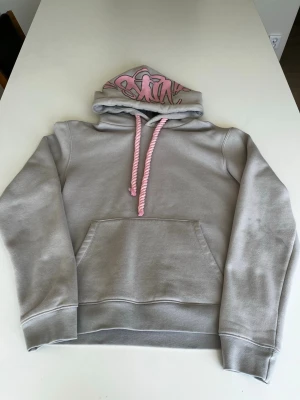 Syna world hoodie - Säljer en grå hoodie med stor ficka framtill och huva. Huvan har rosa graffiti-tryck på insidan och matchande rosa/vita snören. Hoodien har ribbade muddar vid ärmar och nederkant. Perfekt för en chill och avslappnad stil.
