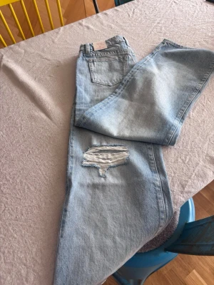 Ljusblå wide leg jeans Gina Tricot - Säljer ett par ljusblå jeans från Gina Tricot med låg midja och breda ben. Jeansen har en snygg slitning på ena benet och klassiska bakfickor. Perfekta för en avslappnad och trendig look.