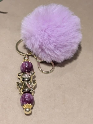 Väsksmycken / keychain - Nyckelring med stor fluffig lila boll och dekoration i form av en guldfärgad björn med vingar samt två lila marmorerade pärlor. Metallring och karbinhake gör den enkel att fästa på väskan eller nycklarna. Perfekt accessoar för att piffa till din stil.
