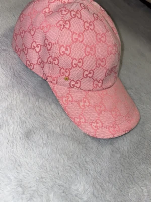 Rosa keps med GG-mönster - Säljer en rosa keps med böjd skärm och helöverdraget GG-mönster i en ljusare och mörkare rosa nyans. Kepsen har klassisk form och är tillverkad i ett tygmaterial. Perfekt för dig som gillar statement-accessoarer.
