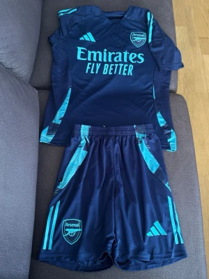 Arsenal mörkblå träningsset - Snyggt Arsenal träningsset från Adidas i mörkblått med turkosa detaljer. Setet består av en t-shirt och shorts med klubbmärke och Adidas-logga. Materialet är lätt och andas, perfekt för träning eller match. Helt ny.