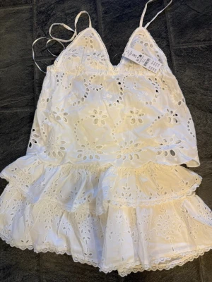 Vitt broderat spetsset med volanger - Säljer ett vitt tvådelat set med broderade blommor och spetsdetaljer. Toppen har smala axelband och v-ringning, kjolen har hög smockad midja och dubbla volanglager. Setet är i luftig bomull och har en romantisk känsla.