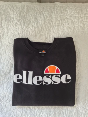 Svart sweatshirt från Ellesse - Svart sweatshirt från Ellesse med stor vit logga och orange/rött tryck på bröstet. Klassisk rund halsringning och långa ärmar. Bra passform