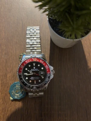 Klocka  - Två ikoniska -klockor: GMT-Master II med svart urtavla, röd och svart vridring samt länk i silverfärgat stål. Datejust med vit urtavla, guldfärgade detaljer och tvåtonad länk i silver och guld. Båda har klassisk rund form och tydliga index.