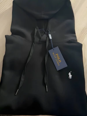 Svart hoodie från Polo Ralph Lauren - Snygg svart hoodie från Polo Ralph Lauren med klassisk huva och dragsnören. Har en liten vit logga broderad på bröstet och en stor magficka framtill. Tillverkad i mjukt material som känns skönt mot huden. Perfekt för en avslappnad och stilren look.