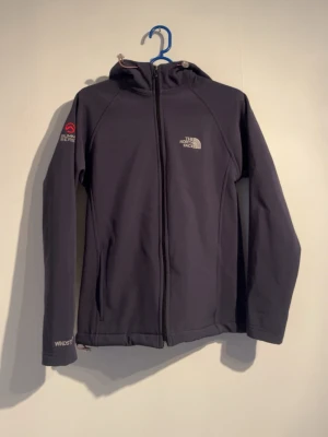 Svart vindjacka från The North Face - Snygg svart vindjacka från The North Face med huva och hel dragkedja. Jackan har WINDSTOPPER-material som skyddar mot vind och är perfekt för blåsiga dagar. Diskreta loggor på bröstet och ärmen samt praktiska fickor framtill.