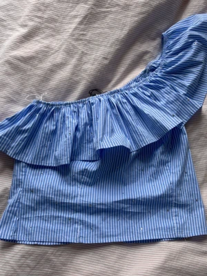 Blå offshoulder topp med volang - Snygg blå offshoulder topp med vita ränder och volangdetalj längs axeln. Toppen är ärmlös och har en luftig passform, perfekt för varma dagar. Tillverkad i bomull för en skön känsla och stilren look.