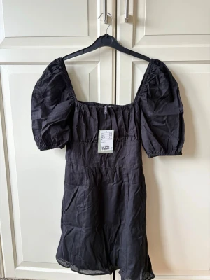 Svart kort klänning med puffärm - Trendig svart kort klänning med puffiga kortärmar och fyrkantig ringning. Tillverkad i ett lätt tyg som ger en luftig känsla. Perfekt för dig som gillar en stilren och modern look.