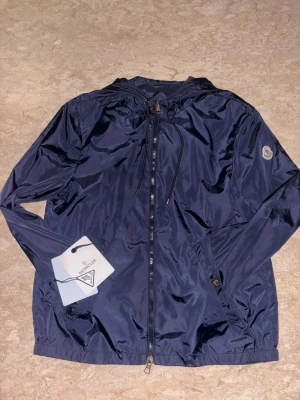 Mörkblå vindjacka från Moncler - Snygg mörkblå vindjacka från Moncler med dragkedja framtill och huva. Använd några få gånger, säljer pågrund utav storleken på jackan. 