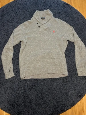 Grå tröja från Polo Ralph Lauren - Snygg grå tröja från Polo Ralph Lauren med hög krage och två knappar. Tröjan har det klassiska röda Polo-logot på bröstet och är tillverkad i mjukt material. Perfekt för en avslappnad men stilren look.