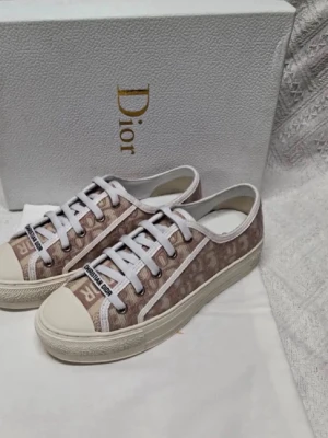 Beige Dior sneakers med monogram - Snygga sneakers från Dior i beige och vitt med klassiskt Dior-monogram över hela skon. Skorna har vita snören, vit sula och tåparti samt detaljer med 'Christian Dior' på plösen. Perfekt för dig som gillar exklusiv och trendig streetstyle.