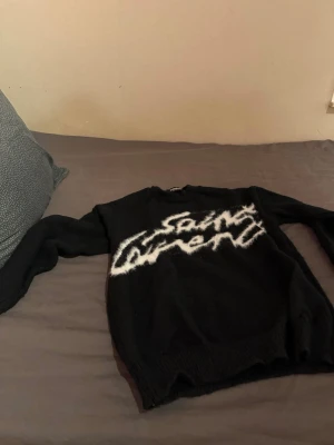 Svart stickad Saint Laurent tröja - Säljer en svart stickad tröja från Saint Laurent med stor vit logga i graffiti-stil över bröstet. Tröjan har lång ärm och rund halsringning. Perfekt för dig som vill ha en statement piece i garderoben.