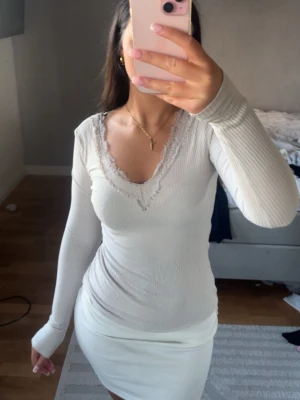 Vit ribbad långärmad topp med spets - Säljer en vit ribbad långärmad topp med v-ringning och fina spetsdetaljer runt halsen. Toppen har en figurnära passform och är perfekt att styla med kjol eller jeans. Superenkel men ändå söt design!