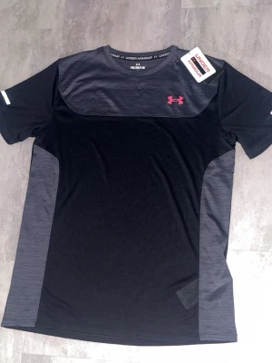 Svart tränings-tshirt Under Armour - Snygg svart och mörkgrå tränings-tshirt från Under Armour med rosa logga på bröstet. Tillverkad i ett lätt och snabbtorkande funktionsmaterial som passar perfekt till gymmet eller löprundan. T-shirten har rund hals och korta ärmar.