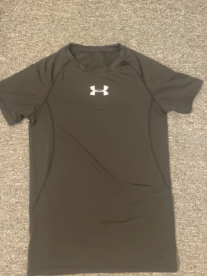 Svart träningströja från Under Armour - Svart kortärmad träningströja från Under Armour med vit logga på bröstet. Tillverkad i ett stretchigt och snabbtorkande funktionsmaterial som passar perfekt för träning och sportaktiviteter. Snygg passform med diskreta sömmar längs sidorna.