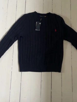 Mörkblå kabelstickad tröja Polo Ralph Lauren - Kabelstickad mörkblå tröja från Polo Ralph Lauren med klassisk röd logga broderad på bröstet. Tröjan har rund halsringning, långa ärmar och ribbade muddar vid ärmslut och nederkant. Perfekt för en stilren och tidlös look. Den har storlek L men är väldigt liten i storlek så passar mera S eller M