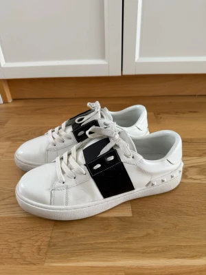 Vita GARAVANI sneakers  - Snygga vita sneakers med bred svart rem över mitten och coola nitar på hälen. Skorna har vita snören och en klassisk låg siluett. Perfekta för dig som vill ha en stilren men ändå edgy look.