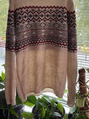 Beige mönstrad stickad tröja Pull&Bear - Stickad tröja från Pull&Bear i beige med vinrött och svart mönster över bröstet och armarna. Tröjan har rund halsringning, långa ärmar och ribbade muddar. Perfekt för dig som gillar klassiska mönster och mjuka material.