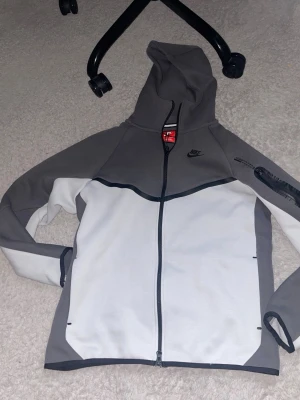 Nike Tech Fleece hoodie grå/silver/vit - Nike Tech Fleece hoodie i grått och vitt med hel dragkedja och huva. Svart Nike-logga på bröstet och svart dragkedja på ärmen. Snygg och sportig design med kontrastpaneler och fickor framtill. Perfekt för en streetwear-look. Undersize så S när de står L, om du köper den nu så kan priset sänkas!!