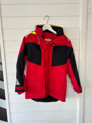 Röd och svart vindjacka från Musto - Snygg röd och svart vindjacka från Musto med gul huva och reflexdetaljer. Jackan har hög krage, dragkedja och flera fickor framtill. Mycket bra skick och inga defekter.