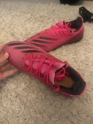 Rosa Adidas fotbollsskor med dobbar - Säljer ett par snygga rosa fotbollsskor från Adidas med svarta ränder på sidan och orange detaljer under sulan. Skorna har snörning och är tillverkade i ett lätt syntetmaterial som ger bra grepp på planen. Perfekta för dig som vill sticka ut på matchen!