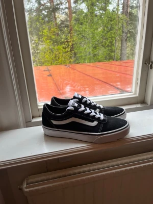 Svarta Vans Old Skool sneakers str 38 - Klassiska Vans Old Skool sneakers i svart med vit sula och vit sidosnöre. Skorna har snörning och är tillverkade i mocka och canvas. Perfekta för en streetwear-look och passar till det mesta. Ikonisk design med Vans-logga på tungan. Bara använda en gång