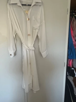 Vit skjortklänning med guldknappar - Stilren vit skjortklänning med långa ärmar, stora guldiga knappar och knytskärp i midjan. Klassisk krage och bröstficka ger en snygg och enkel look. Perfekt för dig som gillar minimalistisk stil.