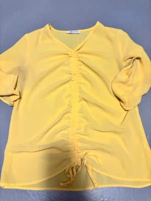 Gul blus med rynk och knytband från Focus - Snygg gul blus i lätt och luftig material med v-ringning och rynkdetalj framtill. Knytband längst ner för passform och trekvartsärmar som ger en avslappnad vibe. Perfekt till jeans eller kjol för en trendig look.