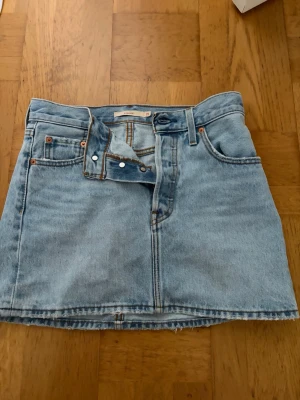 Ljusblå jeanskjol från Levi's - Trendig ljusblå jeanskjol från Levi's med klassisk femficksdesign, rå kant nertill och Levi's ikoniska läderpatch på baksidan! Perfekt till sommaren med superpris! Storlek 25 = xs/s