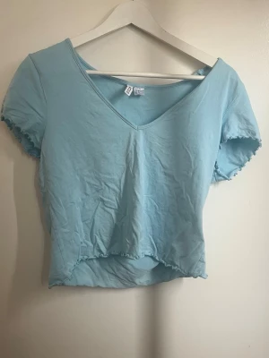 Ljusblå croppad topp med v-ringning - Söt ljusblå croppad topp med v-ringning och korta ärmar. Toppen har vågiga kanter vid ärmslut och nederkant för en extra detalj. Perfekt till jeans eller kjol för en avslappnad look.