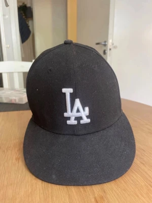 Svart LA Dodgers keps - Säljer en svart keps från New Era med LA Dodgers-logga broderad i vitt framtill. Klassisk böjd skärm och MLB-logga baktill. Perfekt för dig som gillar sportiga accessoarer och streetstyle. Materialet är bomull och kepsen har en strukturerad form.