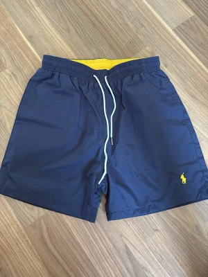 Polo Ralph Lauren badbyxor  - Snygga marinblå badbyxor från Polo Ralph Lauren med gul broderad logga på vänster ben. De har elastisk midja med vit dragsko, gul insida och en bakficka med lock. Perfekta för stranden eller poolen. Passar om du har M OCH S också 