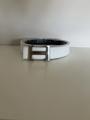 Vitt skärp med silverfärgat H-spänne - Stilrent vitt bälte med ett iögonfallande H-format spänne i silver. Bältet har en klassisk design och är tillverkat i skinn, vilket ger en lyxig känsla. Perfekt accessoar för att lyfta din outfit.