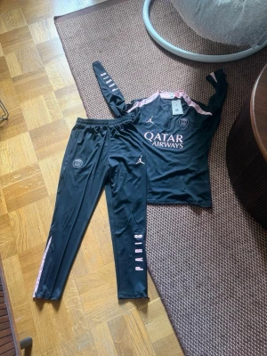 PSG träningsset svart/rosa Jordan - Snyggt träningsset från PSG och Jordan i svart med rosa detaljer. Setet består av långärmad tröja och byxor, båda med PSG-logga och Jordan-logga. Materialet är mjukt och stretchigt, perfekt för träning. Texten 'Qatar Airways' och 'PARIS' syns tydligt på plaggen.