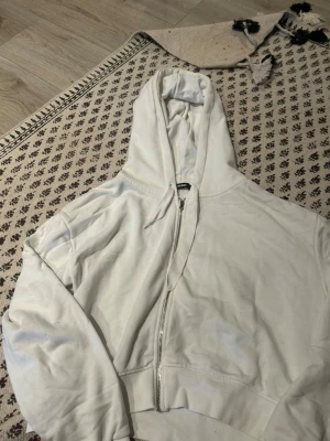 Vit zip-hoodie croppad från Bik Bok - Säljer en vit hoodie från Bik Bok i storlek M. Hoodien har dragkedja framtill, huva med dragsko och ribbade muddar. Perfekt för en chill och sportig look. Materialet är mjukt och skönt bomullstyg.