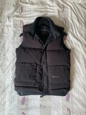 Svart dunväst från Canada Goose - Snygg svart dunväst från Canada Goose med hög krage och två stora fickor framtill. Västen har tryckknappar och dragkedja samt diskret logga på ena fickan. Perfekt för lager-på-lager och håller dig varm under kyliga dagar. Bara att skriva om du har några funderingar😁