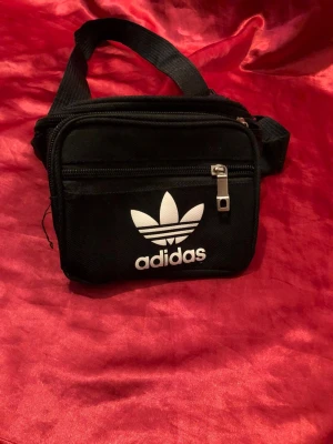 Svart axelväska från Adidas - Snygg svart axelväska från Adidas med klassisk vit logga framtill. Väskan har justerbar axelrem, två dragkedjefack och är tillverkad i slitstark textil. Perfekt för att bära med sig det viktigaste och passar till en sportig eller street-inspirerad stil.