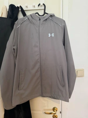 Grå vindjacka från Under Armour - Snygg grå vindjacka från Under Armour med huva och dragkedja framtill. Jackan har två fickor, reflekterande detaljer och stor logga på ryggen. Perfekt för blåsiga dagar och sportiga outfits.