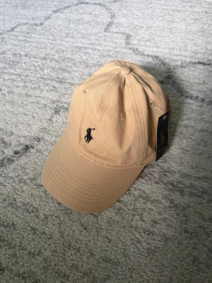 Beige keps från Polo Ralph Lauren - Snygg beige keps från Polo Ralph Lauren med klassisk böjd skärm och broderad logga framtill. Justerbar rem baktill i metall och insida med Polo-mönstrat band. Tillverkad i bomull för en skön och avslappnad look. Passar inte mitt skitiga huvud så köp den haha 