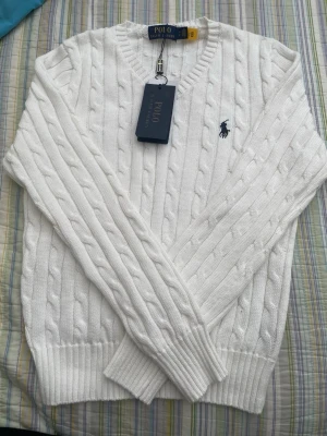 Unik Vit kabelstickad tröja Polo Ralph Lauren - Vit kabelstickad tröja från Polo Ralph Lauren med V-ringning och klassisk marinblå logga broderad på bröstet. Tröjan har ribbstickade muddar och är tillverkad i mjukt material som passar perfekt till en stilren look.