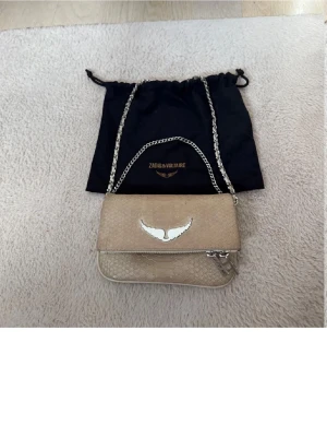 Zadig - Älskar! Snygg beige axelväska från Zadig & Voltaire med silverfärgad kedjerem och ikonisk vinge i metall på framsidan. Väskan har en stilren, rektangulär form och är tillverkad i ett material med ormmönstrad struktur. Perfekt för dig som gillar trendiga accessoarer.