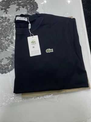 Svart t-shirt från Lacoste - Snygg svart t-shirt från Lacoste med klassisk krokodillogga på bröstet. T-shirten har rund halsringning och korta ärmar. Tillverkad i mjuk bomull för en skön känsla och stilren look. Vi kan snacka om priset dm 