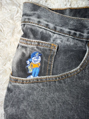 Grå baggy jeans med brodyr - Säljer ett par grå tvättade baggy jeans från Polar skate co med cool broderad figur i blått och gult på fickan. Jeansen har raka ben, hög midja och kontrastsömmar i orange. Perfekta för en avslappnad och trendig streetwear-look.