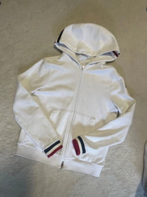 Vit Moncler hoodie med dragkedja - Snygg vit hoodie från Moncler med dragkedja framtill och fickor. Huva och muddar har ränder i blått och rött för en sportig look. Moncler-logga på ärmen. Tillverkad i mjuk bomull som känns skön mot huden.