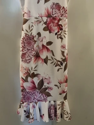Blommig volangklänning från Boohoo - Supersöt vit klänning från Boohoo med stora blommor i rosa och lila toner samt gröna blad. Klänningen har volang nertill och en rak passform. Perfekt för sommarens alla tillfällen!