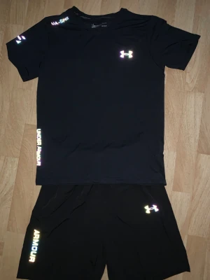 Svart träningsset från Under Armour - Säljer ett svart träningsset från Under Armour med t-shirt och shorts. Setet har reflekterande loggor och textdetaljer i vitt och regnbågsskimrande färg. Materialet är lätt och snabbtorkande, perfekt för träning och sport.